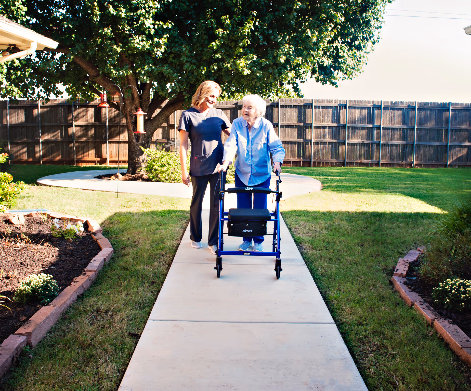 An elderly woman using a walker is accompanied by a caregiver walking together on a paved path in a sunny garden with green grass, flower beds, and a large tree in the background.