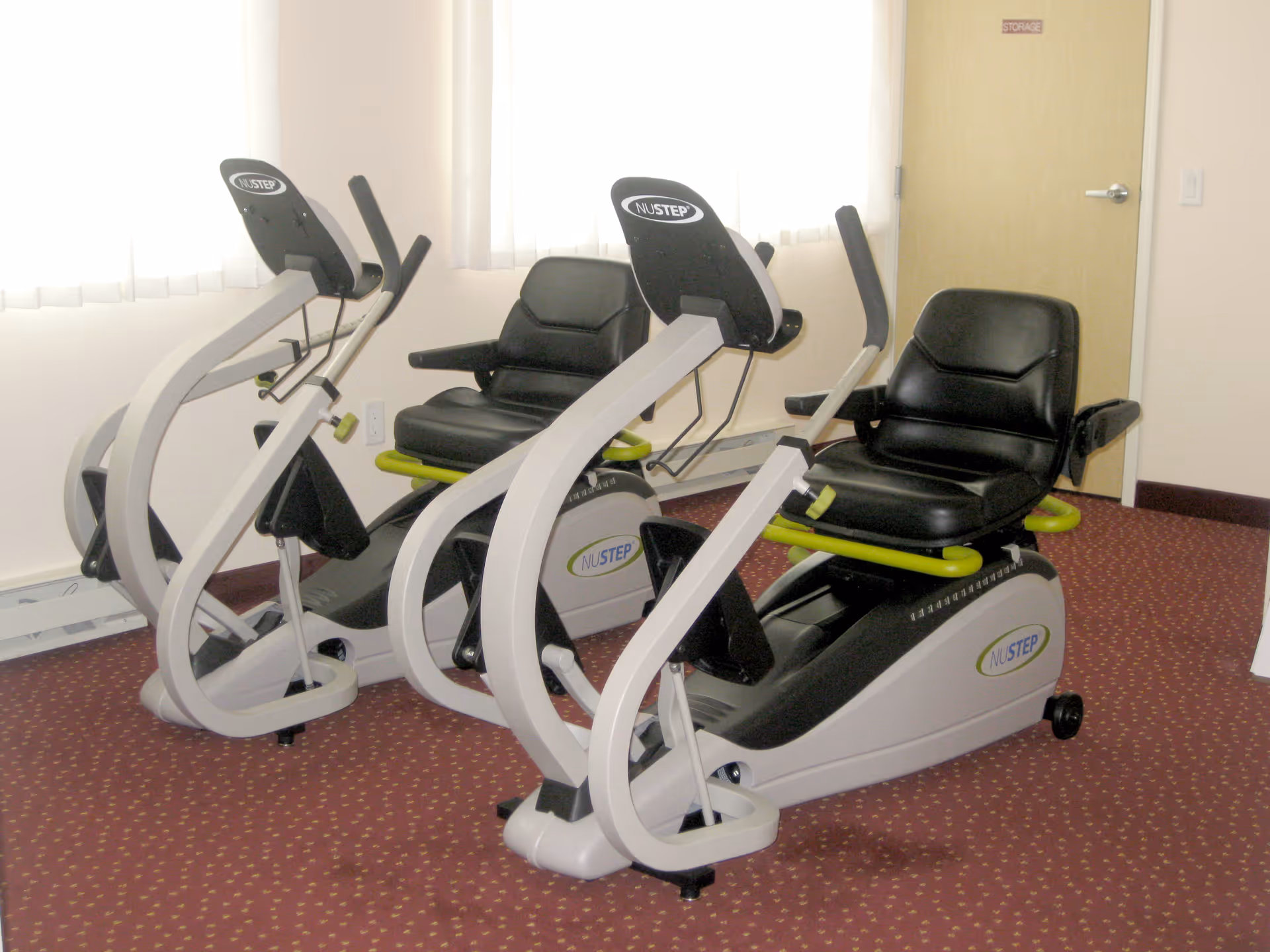 Two NuStep recumbent cross trainers with black seats and arm handles placed on a red carpeted floor in a room with beige walls and a closed wooden door labeled 'Storage'.