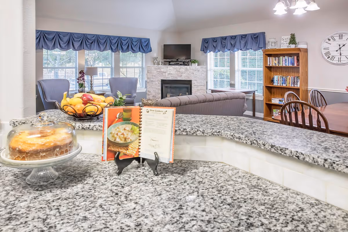 View from a granite kitchen counter looking into a living area with a fireplace, a TV mounted above it, two blue armchairs near windows with blue valances, a wooden bookshelf filled with books, a dining table with chairs, and a cake under a glass dome on the counter along with a bowl of fruit and an open cookbook on a stand.