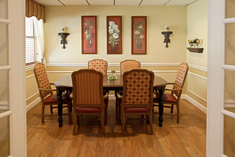 Dining room with a wooden table surrounded by six upholstered chairs, floral wall art, and decorative wall sconces.