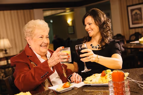 An elderly woman and a younger woman smiling and toasting with drinks over a table with a cheese and fruit platter.