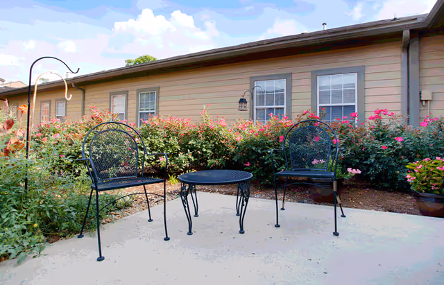 Outdoor patio area with two black metal chairs and a matching round table on a concrete surface, surrounded by flowering bushes and plants in front of a beige building with windows.