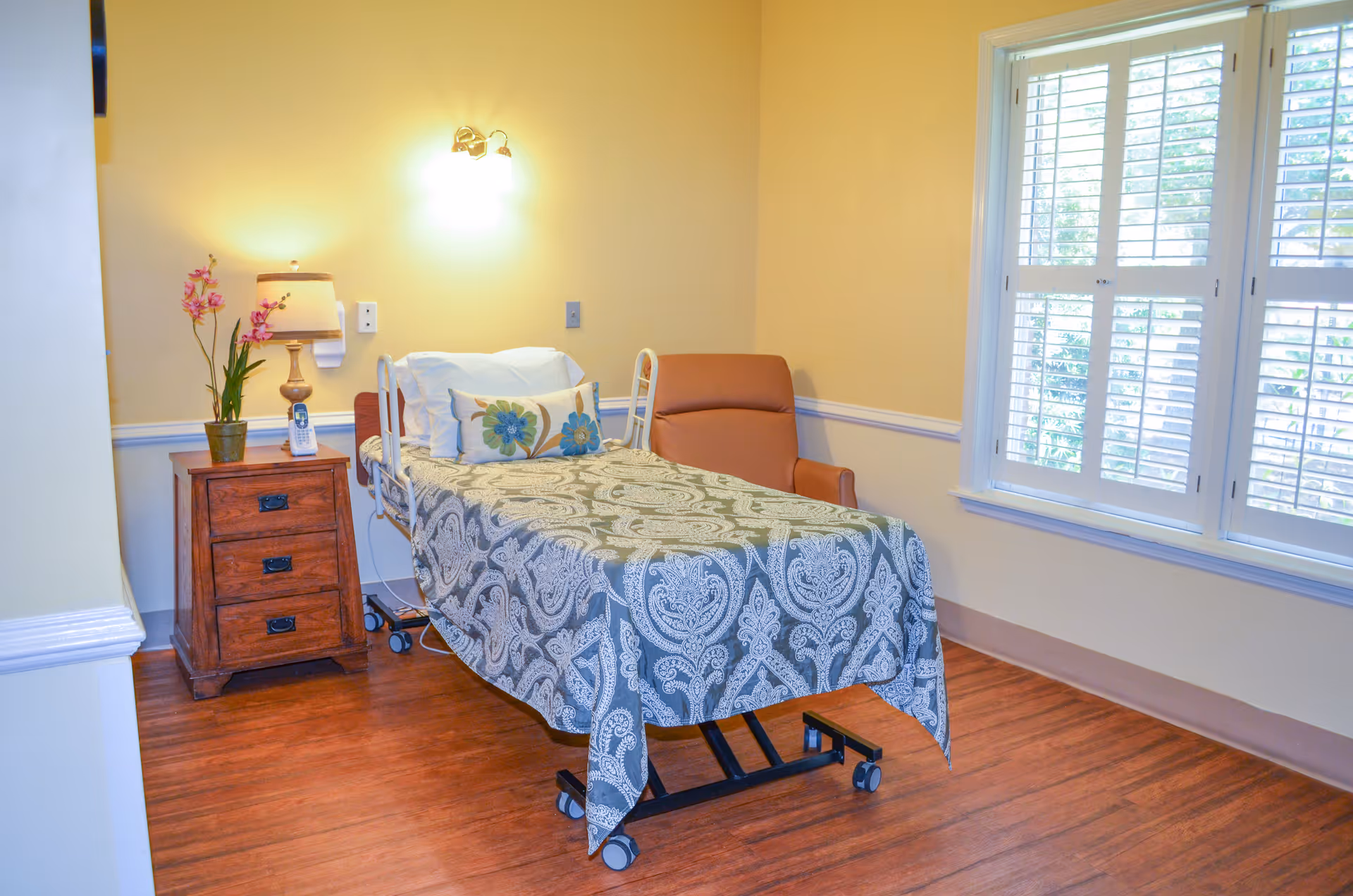 A small bedroom with a single hospital-style bed covered with a patterned bedspread and a floral pillow. Next to the bed is a wooden nightstand with a lamp, a potted orchid, and a cordless phone. There is a comfortable armchair beside the bed and a window with white plantation shutters letting in natural light. The walls are painted a soft yellow and the floor is wood.