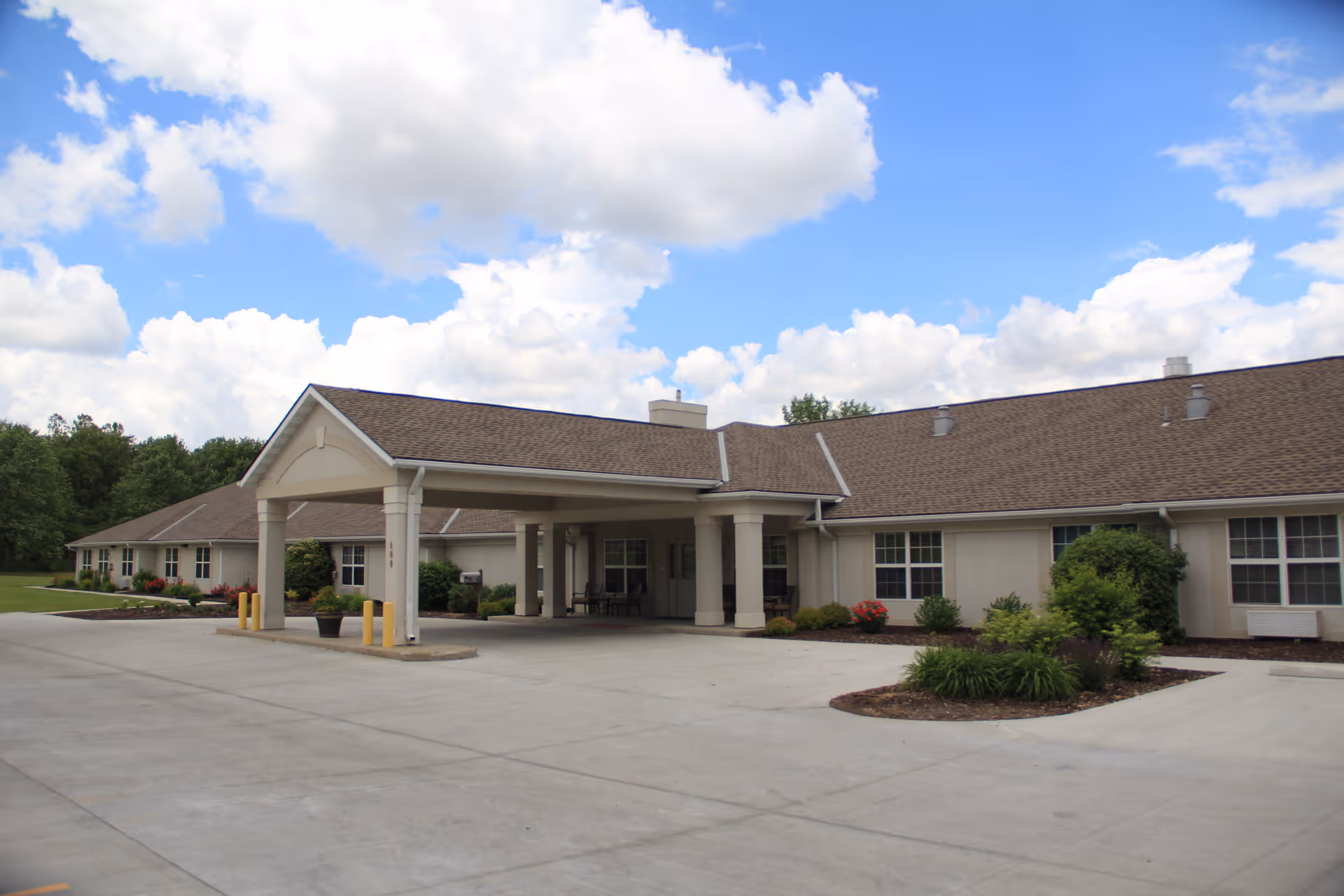 Exterior view of a single-story senior living facility building with a covered entrance, beige walls, brown shingled roof, and landscaped greenery under a partly cloudy blue sky.