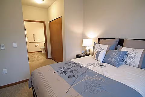 A bedroom with a neatly made bed featuring floral-patterned bedding and multiple pillows. To the right of the bed is a nightstand with a lamp. The room has beige walls and carpeted floor. An open doorway reveals a bathroom with a toilet and grab bars visible.