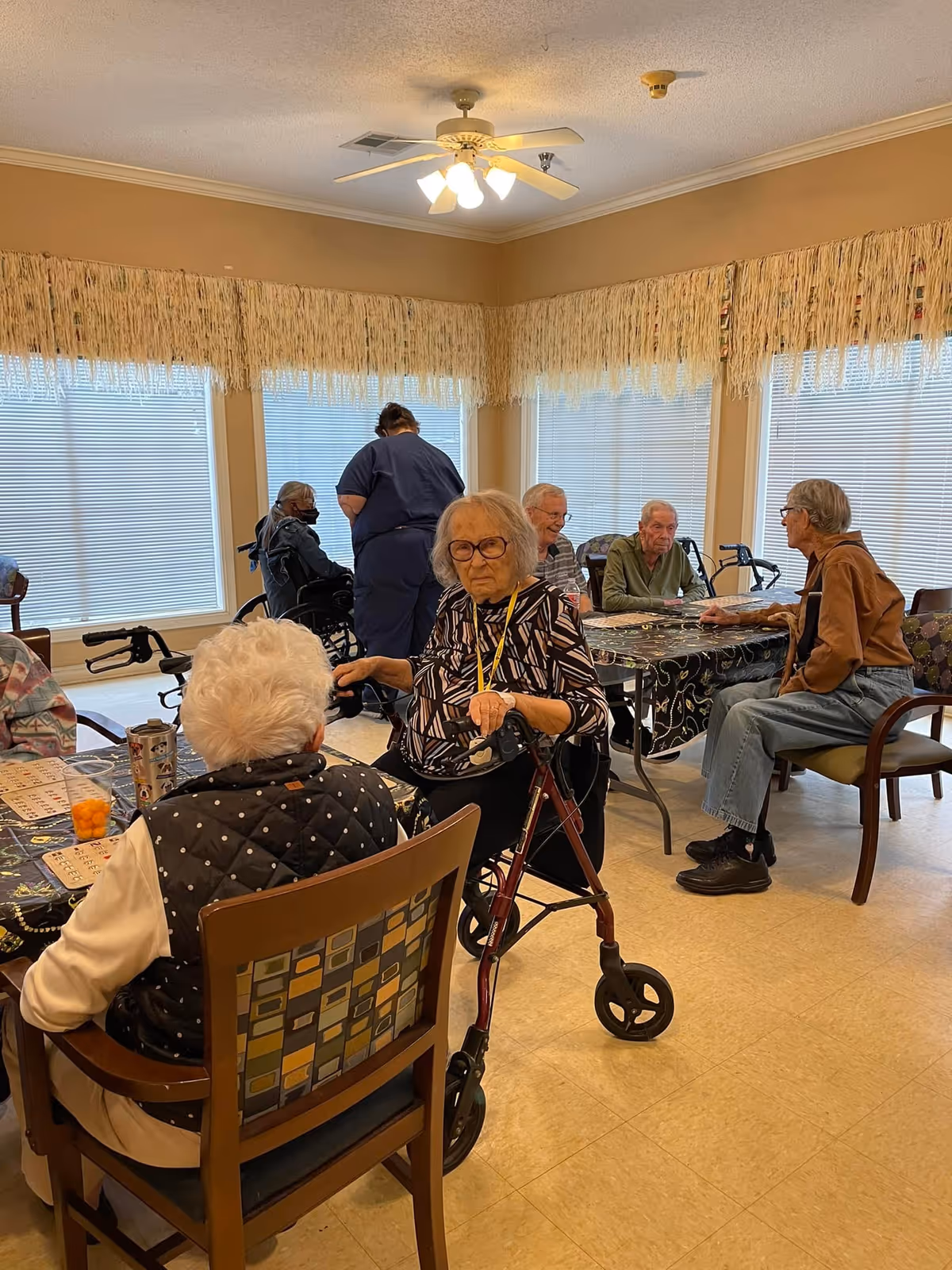 A group of elderly people sitting and standing around tables in a well-lit room with large windows covered by blinds and fringed valances. Some individuals are using walkers and wheelchairs, and a caregiver in blue scrubs is assisting one of the residents. The room has beige walls and a ceiling fan with lights.