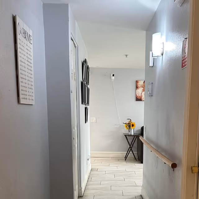 A hallway in a care home with light gray walls and tiled floor. On the left wall, there is a decorative sign with text and several framed pictures. On the right wall, there is a handrail and a wall-mounted light fixture. At the end of the hallway, there is a small table with a vase of sunflowers and a piece of wall art above it.