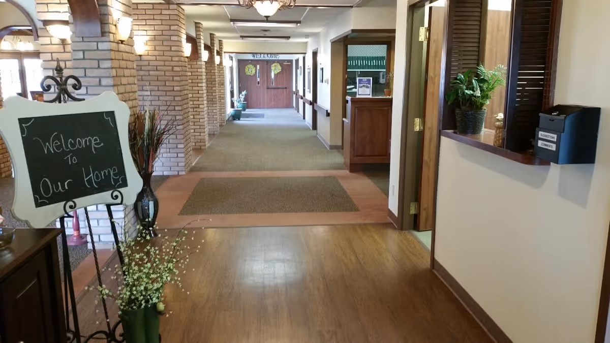 Interior hallway of an assisted living facility with wooden floors and brick columns. A chalkboard sign on a stand reads 'Welcome to Our Home'. There are plants and a reception window on the right side. The hallway extends toward double doors at the far end.
