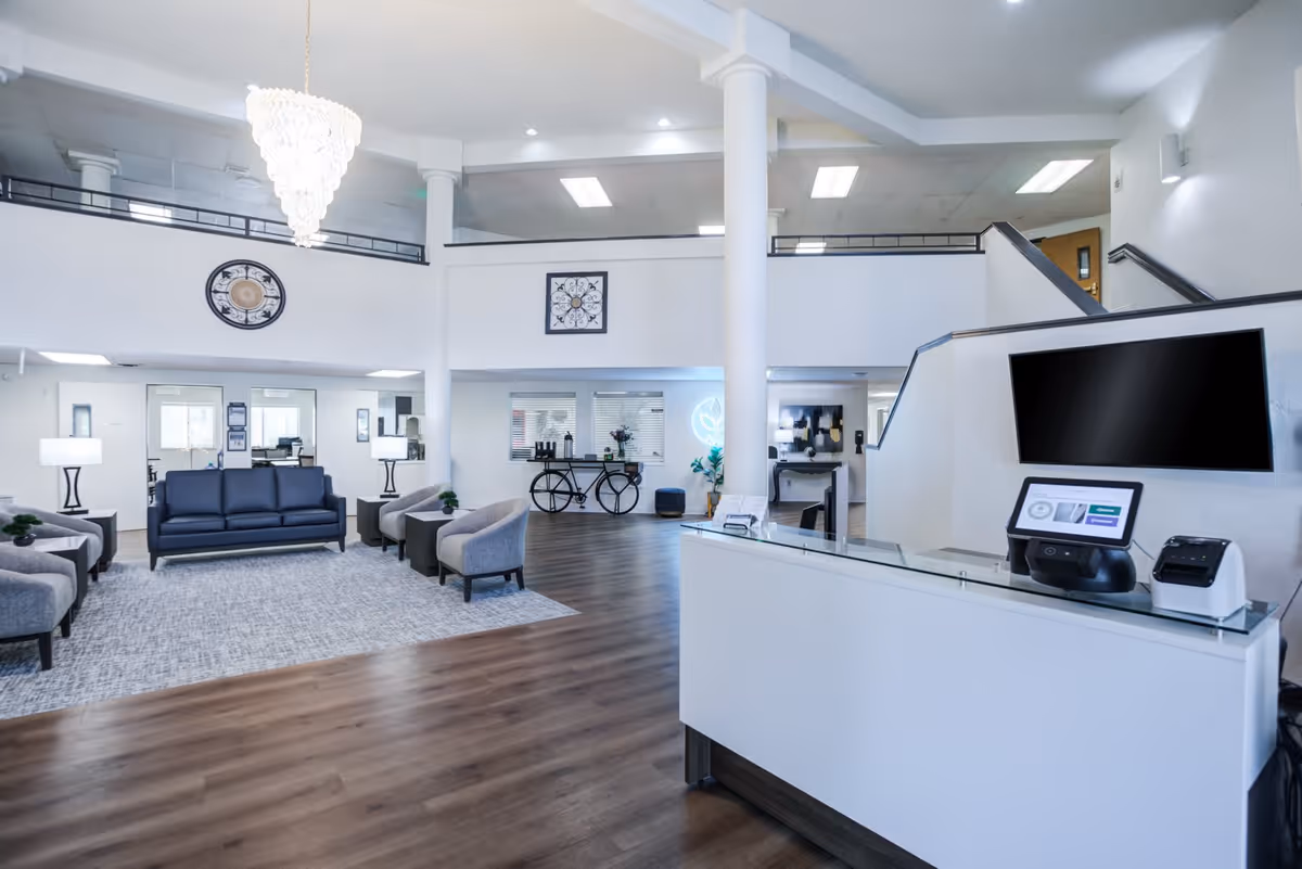 Spacious and modern senior living facility lobby with a high ceiling, white walls, and wooden flooring. The area features a reception desk with a computer and printer, a seating area with a dark blue sofa and gray armchairs arranged around a light gray rug, two table lamps, decorative wall clocks, and a chandelier hanging from the ceiling. There is also a decorative bicycle and plants near the back wall.