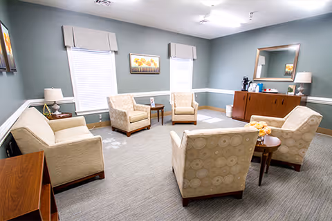 A cozy sitting area with beige upholstered chairs and a matching sofa arranged around small wooden tables. The room has light gray walls with white trim, two windows with white blinds and valances, a wooden sideboard with a lamp and decorative items, and a large mirror above it. The floor is carpeted in a neutral tone.