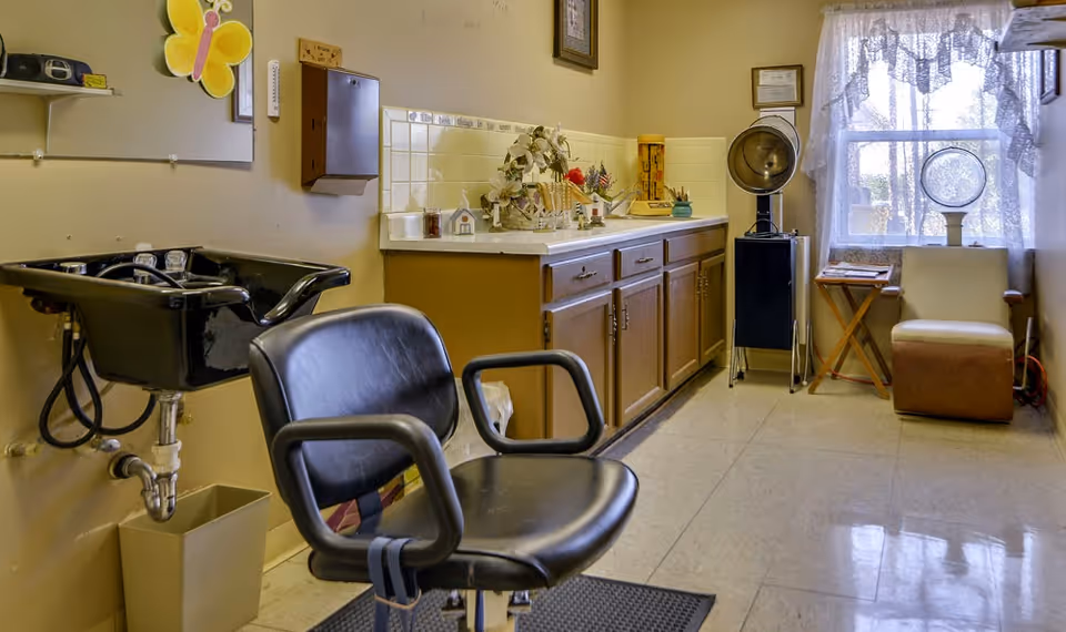 Interior of a small hair salon area in a senior living facility with a black salon chair and black wash basin on the left, a countertop with cabinets underneath along the back wall, a vintage hair dryer on a stand near a window with lace curtains, and a beige chair with a small wooden table beside it.