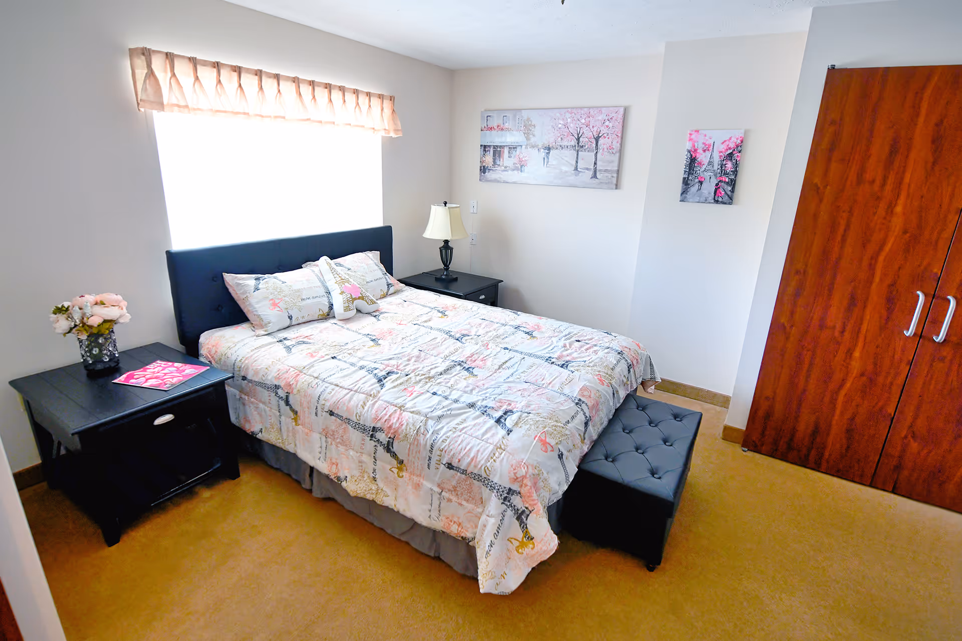 A bright bedroom with a large window covered by a beige valance. The room features a bed with a patterned quilt and matching pillows, a black tufted headboard, a black nightstand with a lamp, a small black ottoman at the foot of the bed, a vase with flowers on another black nightstand, and two framed pictures on the walls. There is a wooden wardrobe with silver handles on the right side.