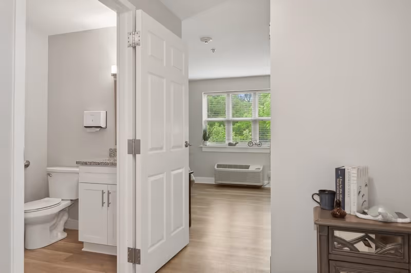 Interior view of an apartment featuring an open bathroom with a toilet and vanity, a bright main room with a window and wall air conditioner, and a small table with books and decor.