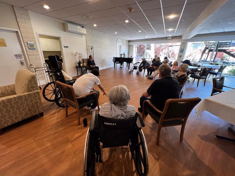 A group of elderly individuals seated in a circle in a spacious room with wooden flooring and large windows letting in natural light. Some people are in wheelchairs, and a staff member is engaging with the group. There is a piano in the background and comfortable chairs arranged around the room.