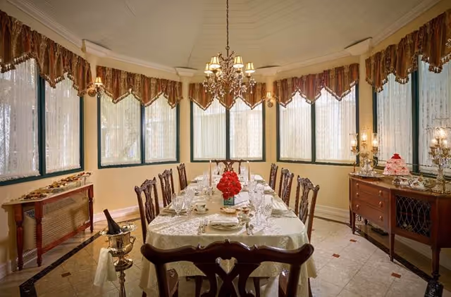 A formal dining room with a long table set for a meal, featuring a white tablecloth, glassware, plates, and a centerpiece of red flowers. The room has multiple tall windows with lace curtains and decorative valances. There is a chandelier hanging above the table, and side tables with decorative items including a cake and lamps.
