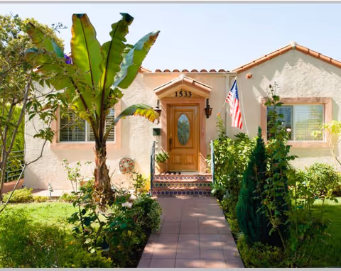 Front exterior view of a single-story house with a tiled roof, a wooden front door with an oval glass panel, the number 1533 above the door, an American flag mounted on the right side, and a walkway lined with green plants and trees leading up to the entrance.