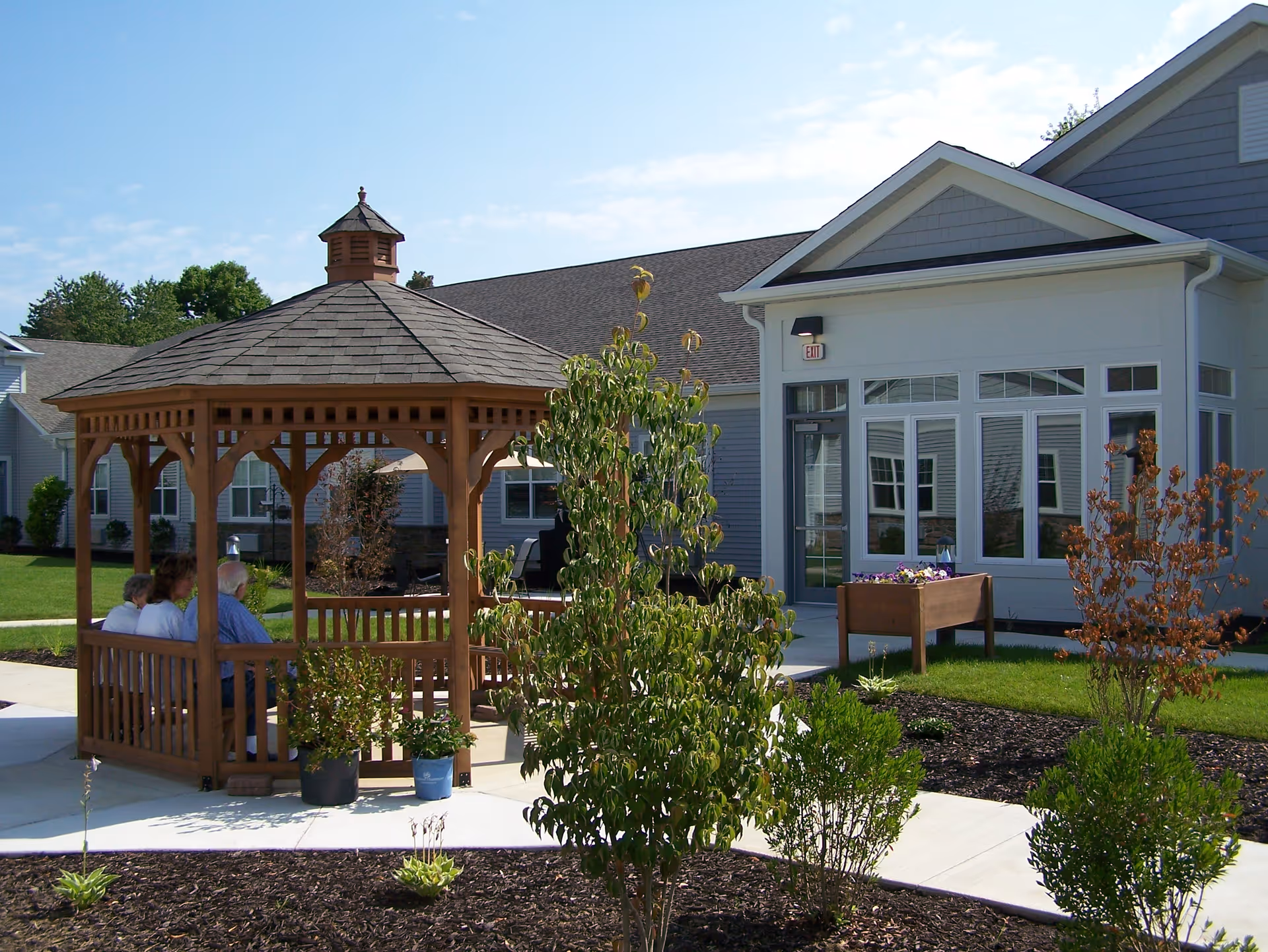 Outdoor scene at North Woods Village of Kalamazoo featuring a wooden gazebo with three elderly people sitting inside. The gazebo is surrounded by a well-maintained garden with small trees, shrubs, and flower beds. In the background, there is a light gray building with large windows and a door marked with an exit sign. The sky is clear with some clouds.