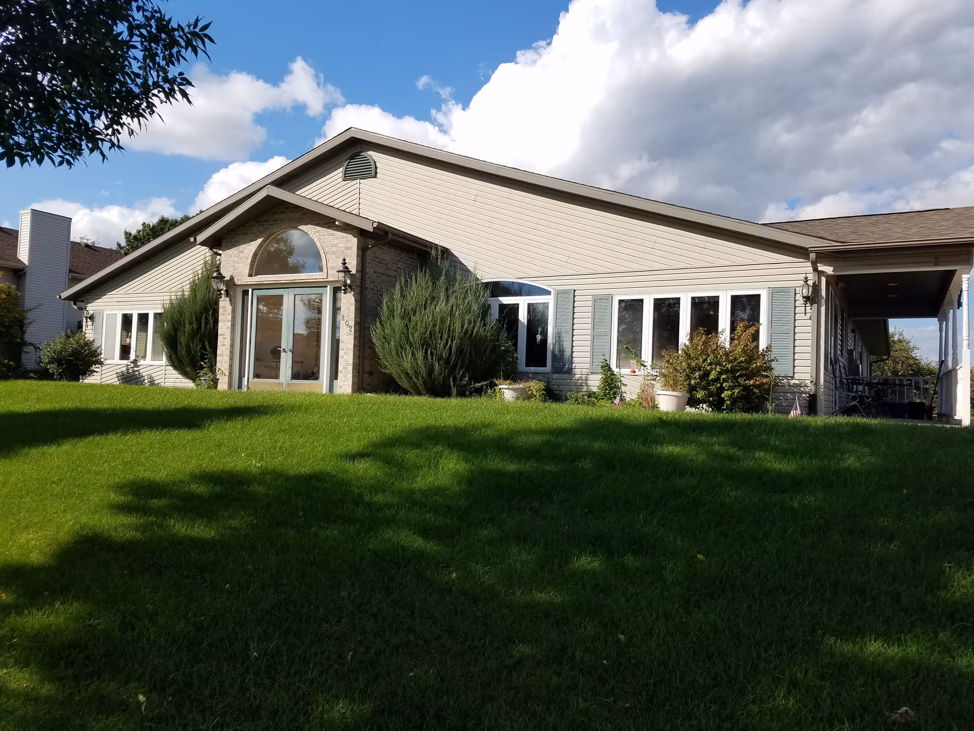 Exterior view of a single-story building with beige siding and a brick entrance. The building has multiple windows with blue shutters and is surrounded by green grass and shrubs under a partly cloudy sky.
