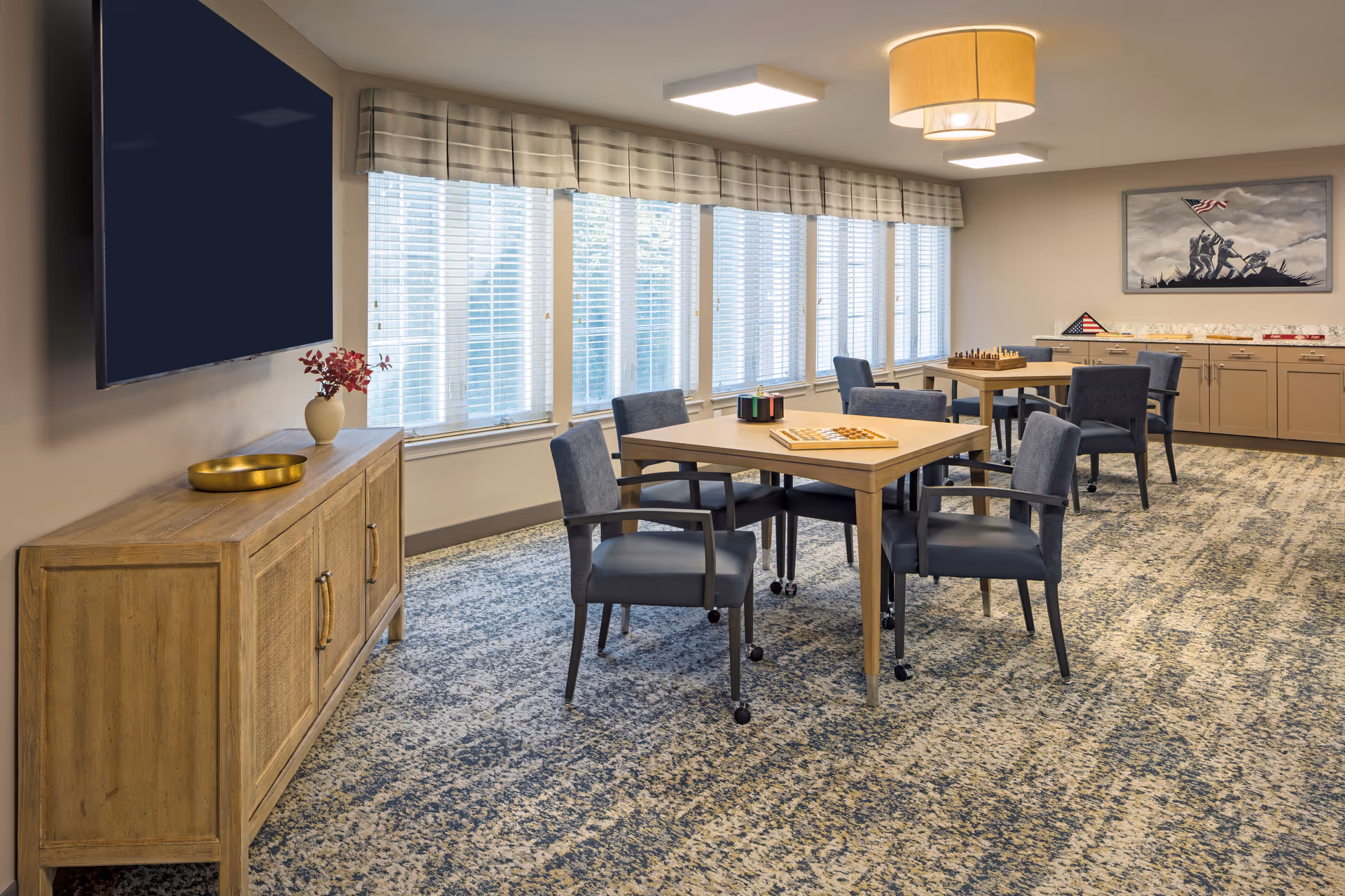 Bright activity/dining room with several tables and chairs, a wall-mounted TV, large windows, and cabinets with games and decor.