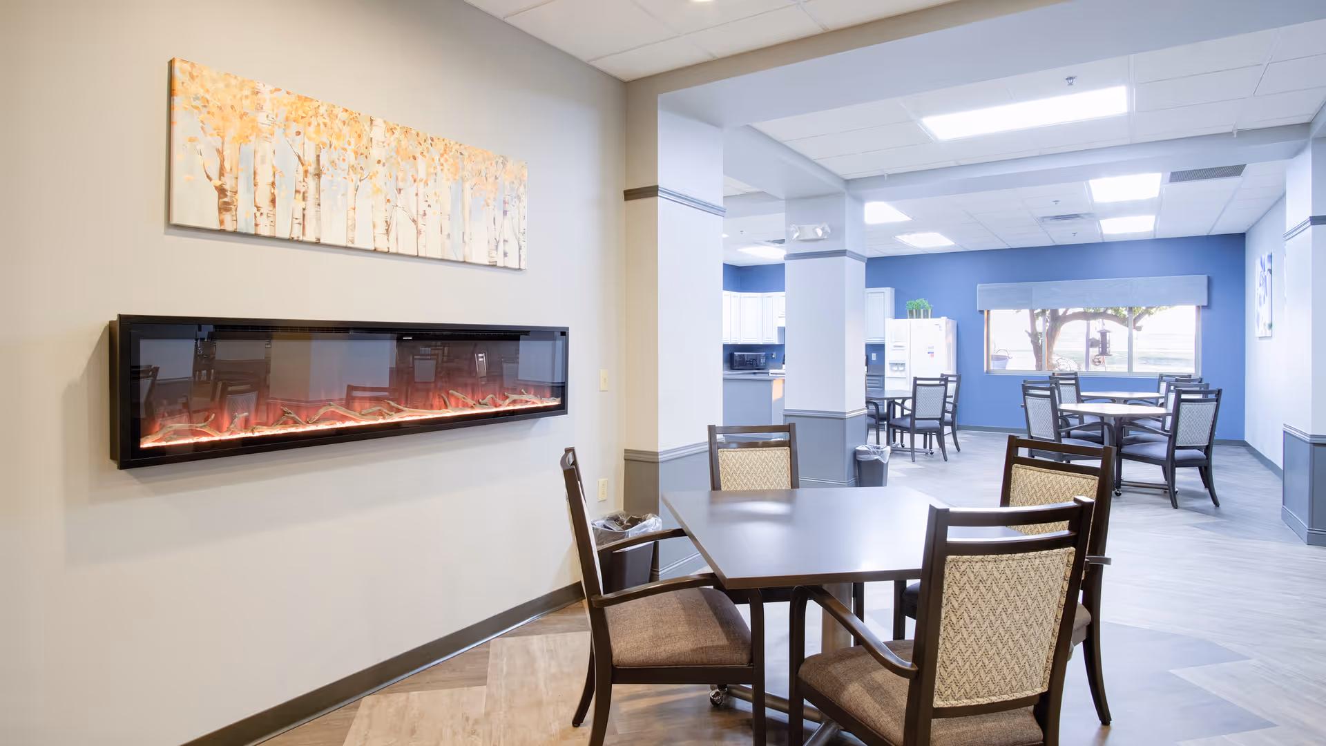 A bright and spacious dining area in a senior living facility featuring multiple square tables with cushioned chairs. On the left wall, there is a modern electric fireplace beneath a painting of birch trees with golden leaves. The room has light-colored walls and flooring, with a blue accent wall in the background near a large window letting in natural light. The kitchen area is visible in the background with white cabinets and appliances.
