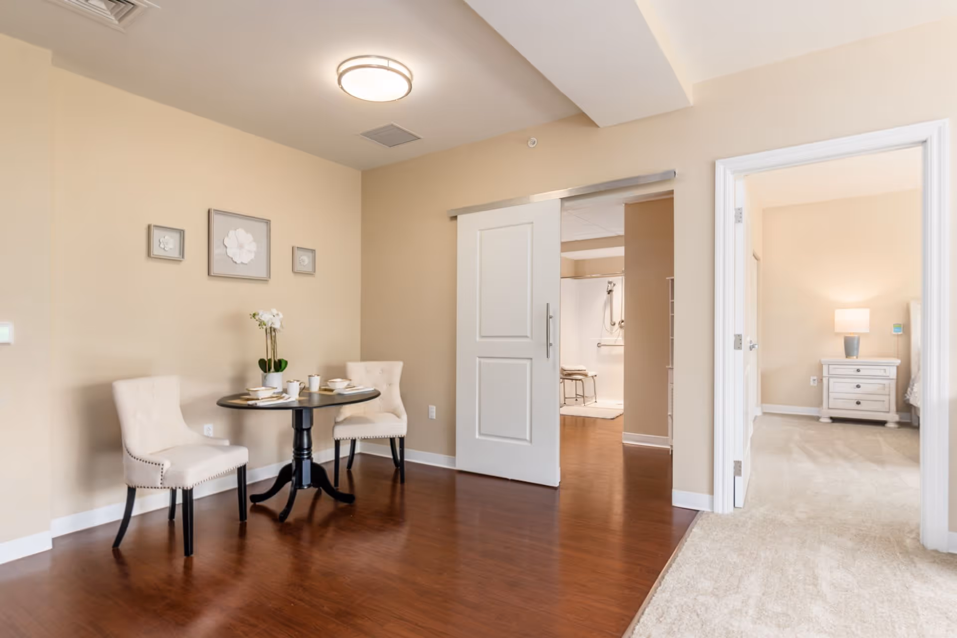 A small dining area with a round black table set for two with white chairs, a flower vase centerpiece, and beige walls decorated with three framed flower artworks. To the right, there is an open doorway leading to a carpeted bedroom with a nightstand and lamp, and a sliding door leading to a bathroom with grab bars and a shower chair.