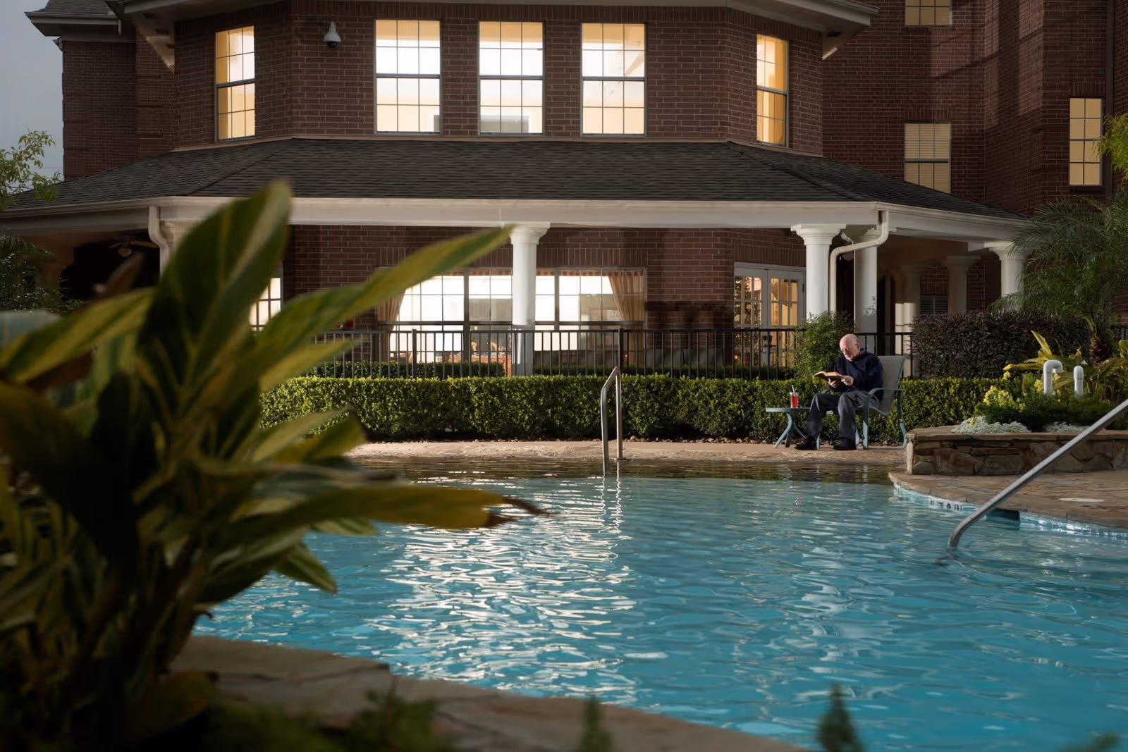 An elderly man sitting on a chair by a swimming pool at night, reading a book with a drink on a small table beside him. The background shows a brick building with lit windows and a covered patio area surrounded by greenery.