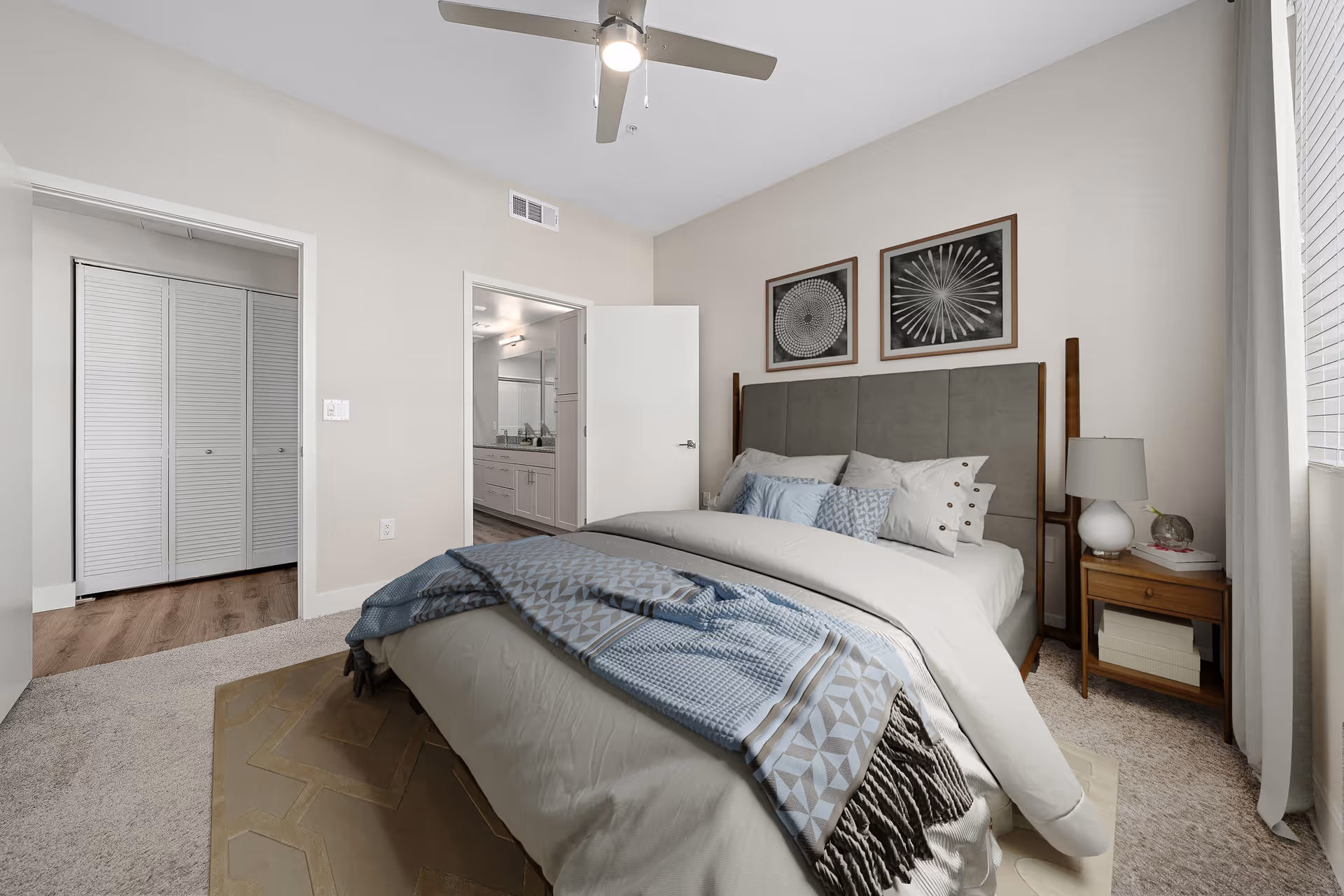 A modern bedroom with a large bed covered in white and blue bedding, a wooden nightstand with a lamp and decorative items, two framed abstract artworks above the headboard, a ceiling fan, and an open door leading to a bathroom and closet area.