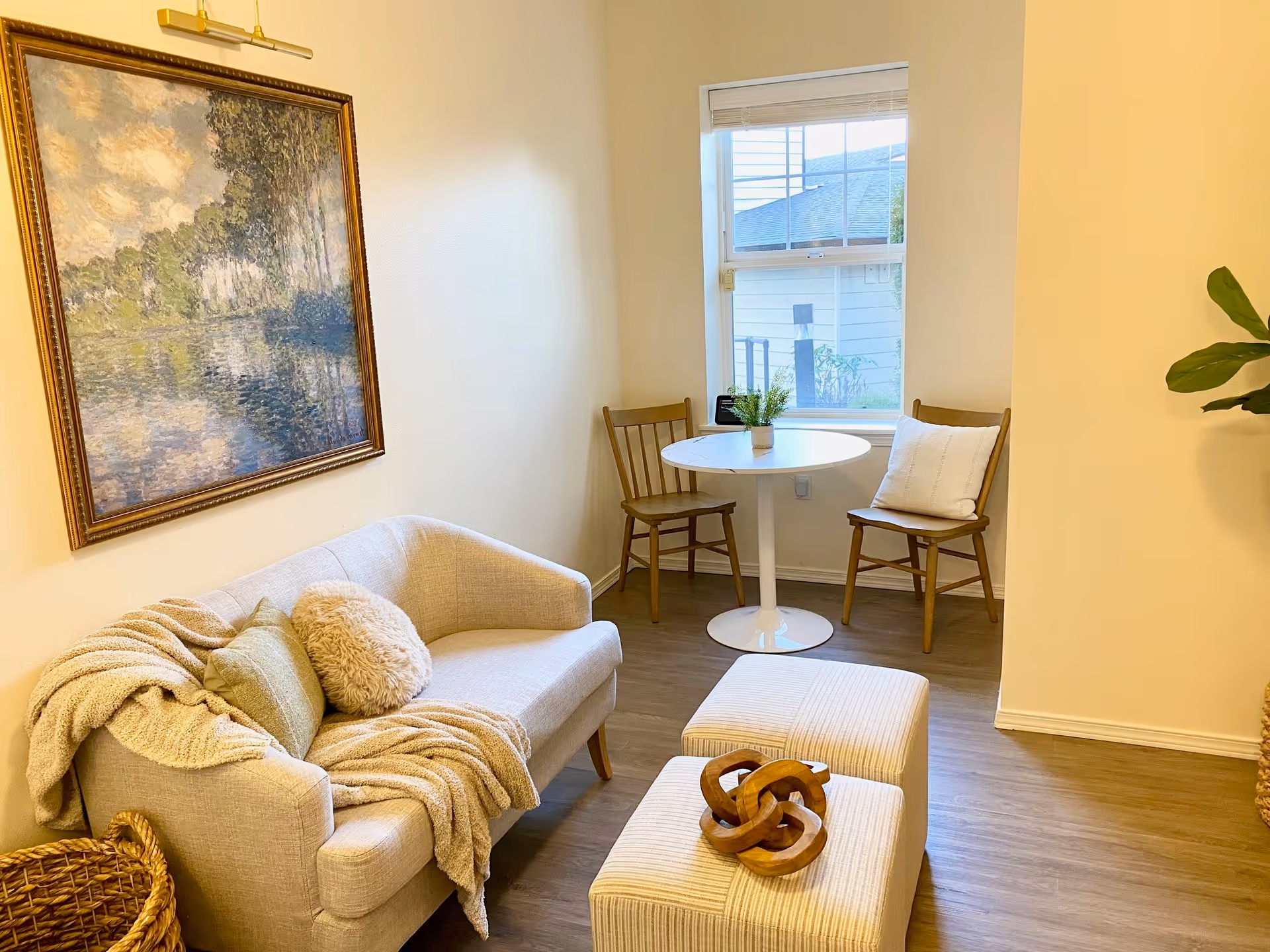 A cozy living room area with a beige loveseat adorned with a throw blanket and pillows. In front of the loveseat are two cushioned ottomans with a decorative wooden sculpture on top. A round white table with two wooden chairs is positioned near a window with a view of a neighboring building. A large framed painting hangs on the wall above the loveseat, and a green plant is partially visible on the right side.