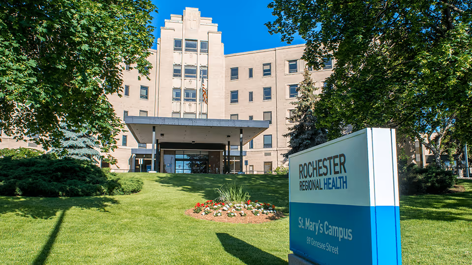 Exterior view of a large multi-story beige building with a covered entrance, surrounded by green trees and a well-maintained lawn with a flower bed. A sign in the foreground reads 'Rochester Regional Health St. Mary's Campus 89 Genesee Street' under a clear blue sky.