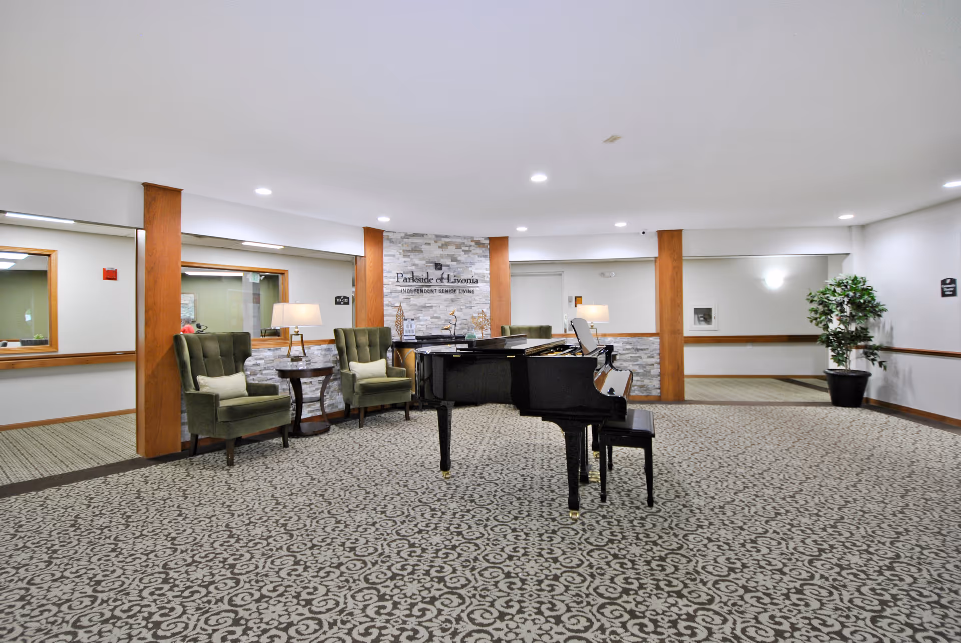 Bright senior living facility common area with a grand piano, two green armchairs, patterned carpet, and a stone accent wall reading "Parkside of Livonia."