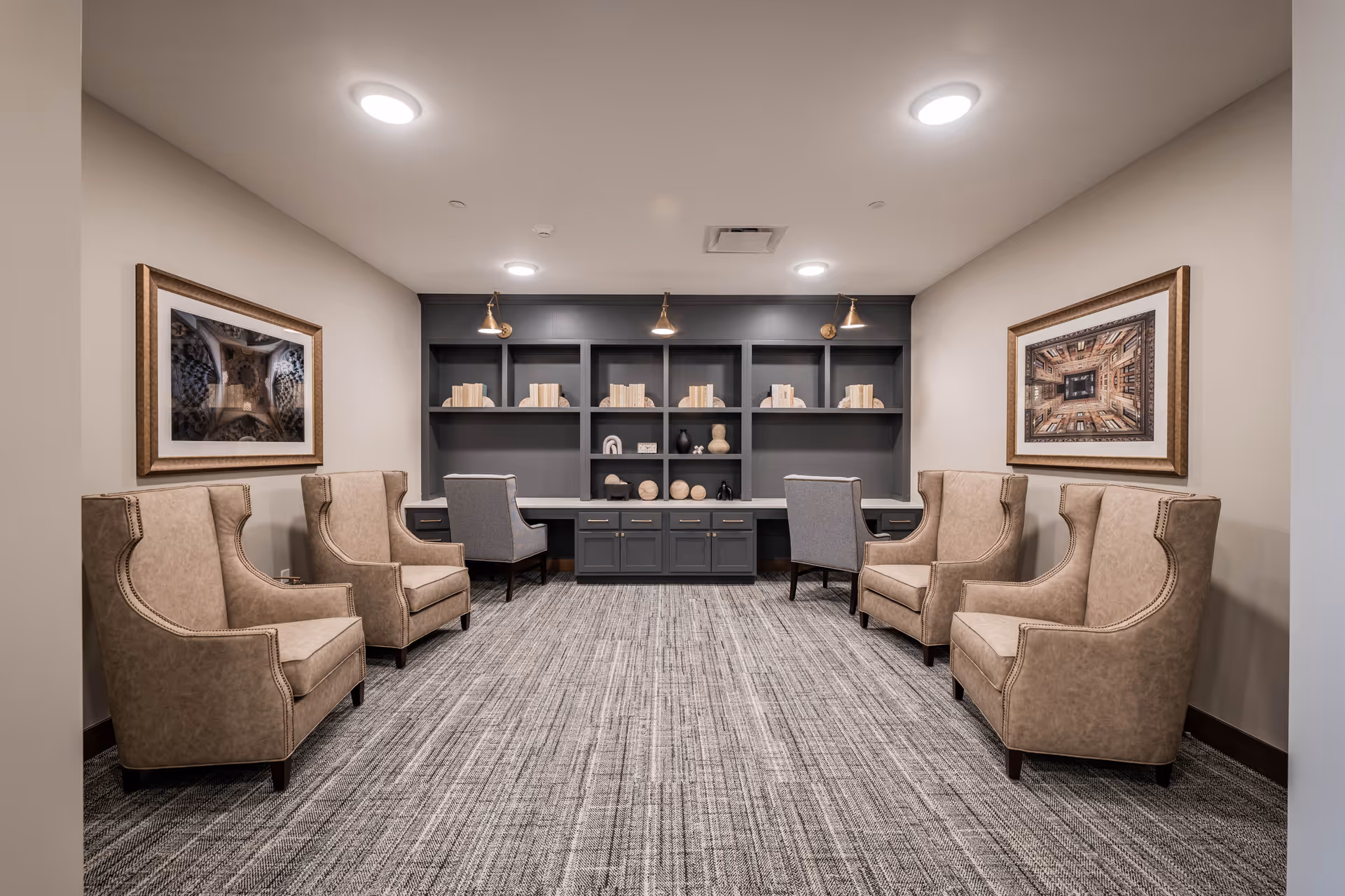 A quiet seating area in a senior living facility featuring six beige upholstered armchairs arranged in two rows facing each other, with two gray chairs positioned at a built-in desk with shelves filled with decorative items and books. The walls are adorned with framed artwork, and the room has a neutral color palette with carpeted flooring and recessed ceiling lights.