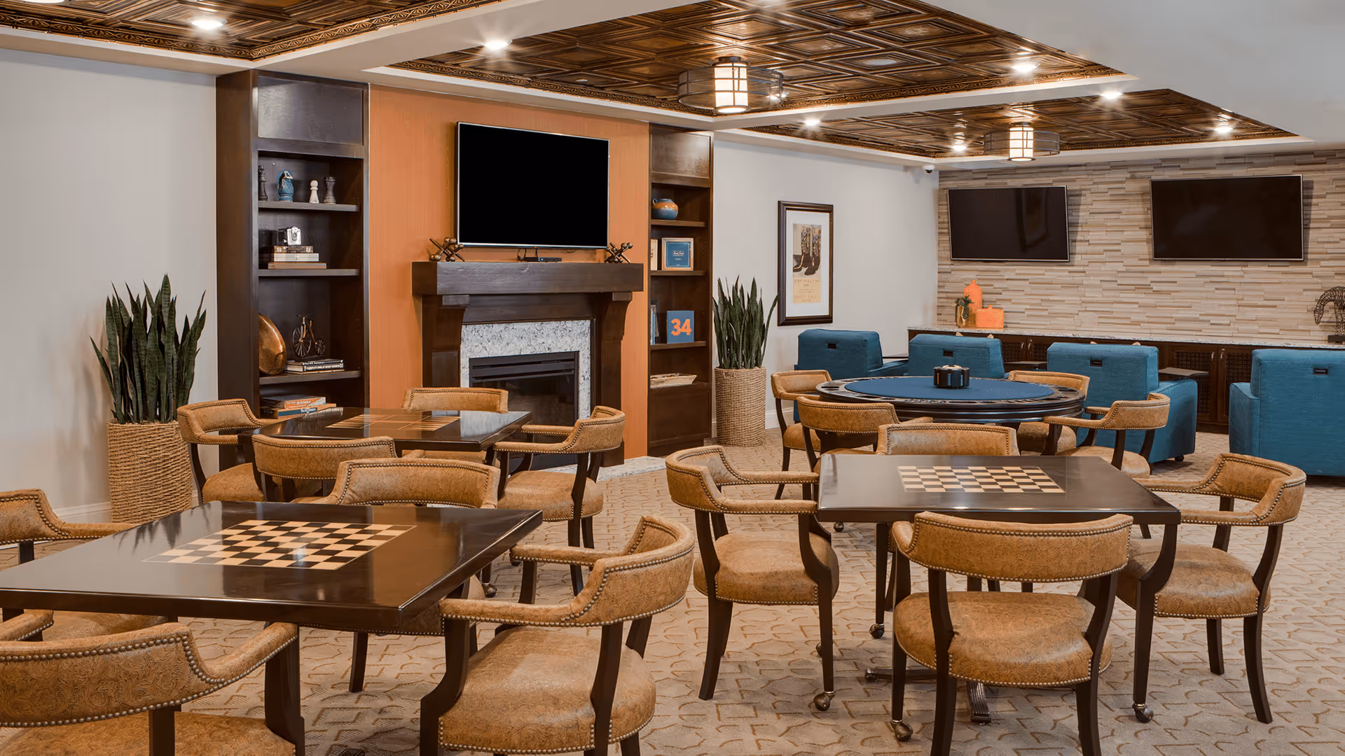 A spacious common room with multiple tables and chairs arranged for games and socializing. The tables have built-in checkerboards. There is a fireplace with a mounted TV above it, built-in shelves on either side, and two additional TVs mounted on a stone accent wall. Comfortable blue armchairs are arranged near the TVs. The room features a decorative wooden ceiling and carpeted floor.