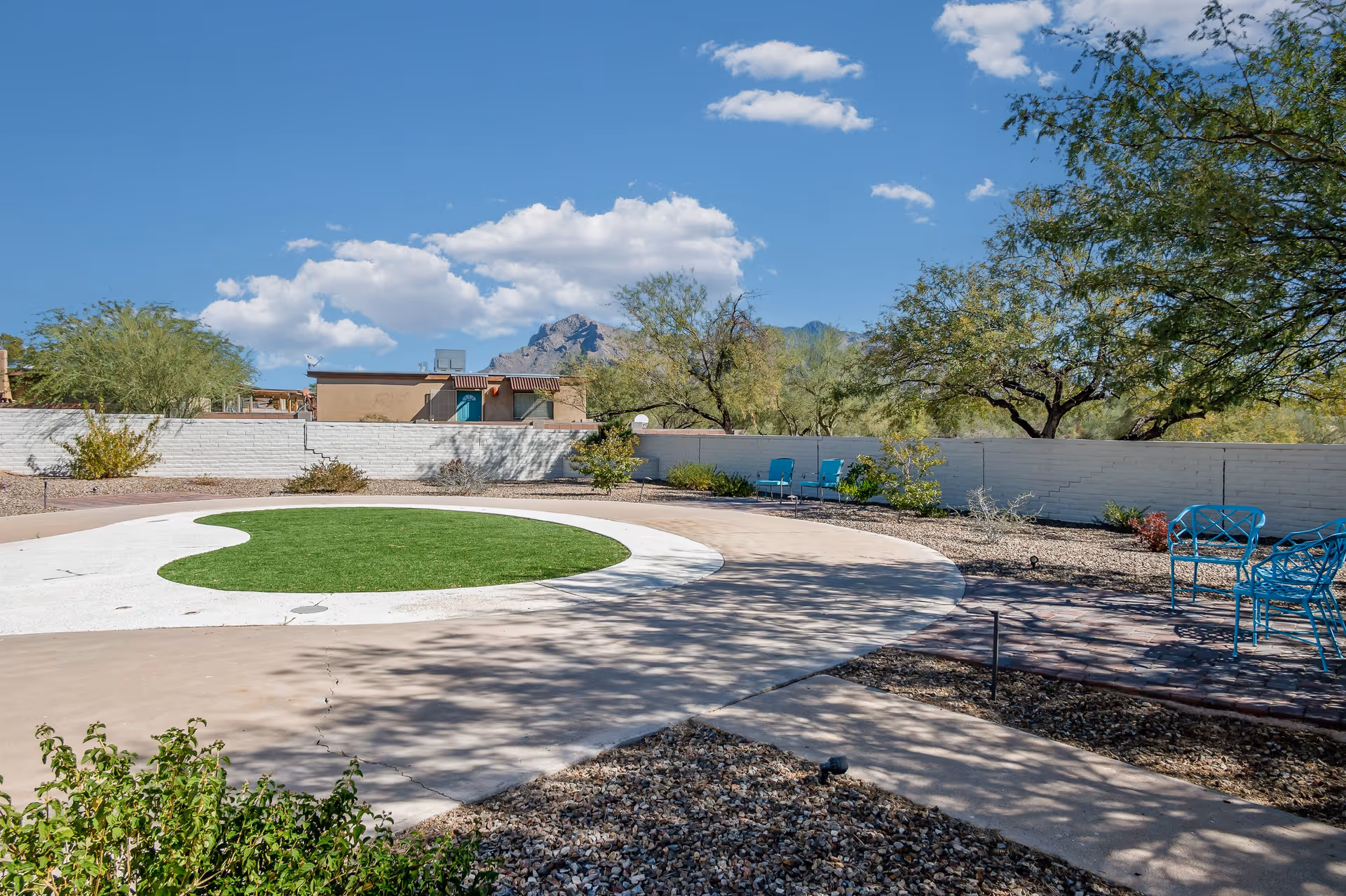 Outdoor area with a circular patch of green grass surrounded by a white concrete border, desert landscaping with gravel and bushes, several blue metal chairs, trees, a white brick wall, and mountains in the background under a blue sky with scattered clouds.