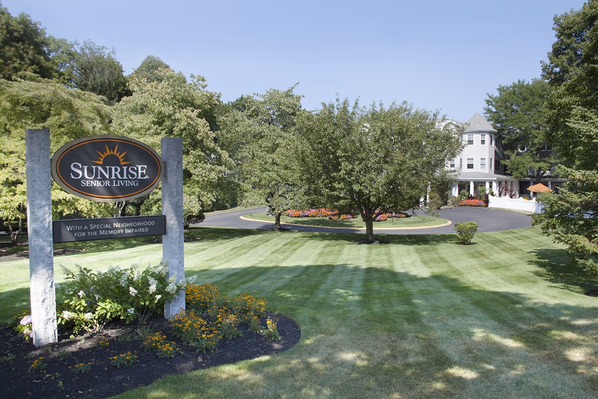 Outdoor view of Sunrise Senior Living facility with a sign in the foreground that reads 'Sunrise Senior Living, With a Special Neighborhood for the Memory Impaired.' The scene includes a well-maintained lawn, trees, flower beds, a circular driveway, and a large building in the background under a clear blue sky.