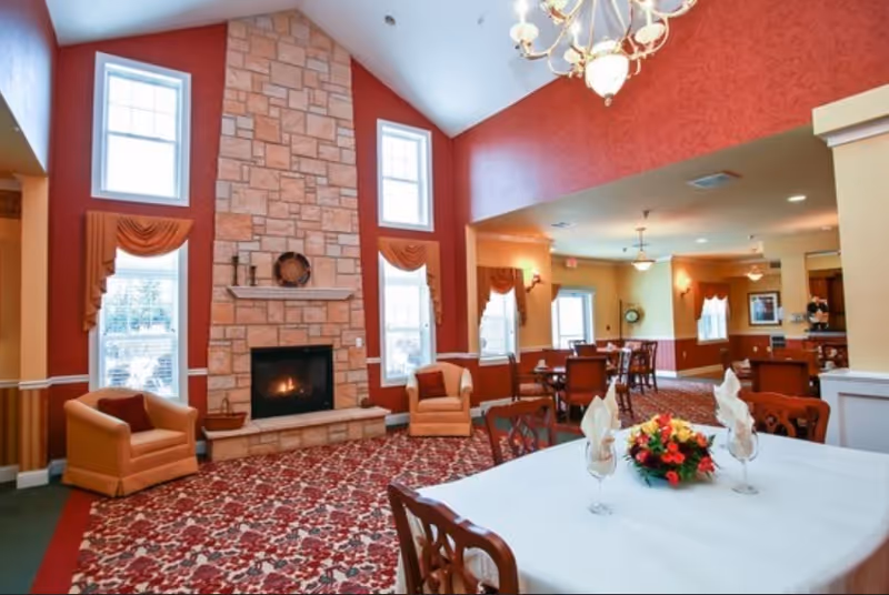 A spacious dining and lounge area with a high ceiling and a large stone fireplace in the center. The walls are painted red and beige, with tall windows adorned with orange drapes. There are comfortable armchairs near the fireplace and several dining tables with chairs, one table set with a white tablecloth, napkins, wine glasses, and a floral centerpiece. A chandelier hangs from the ceiling, and the carpet has a red and beige floral pattern.