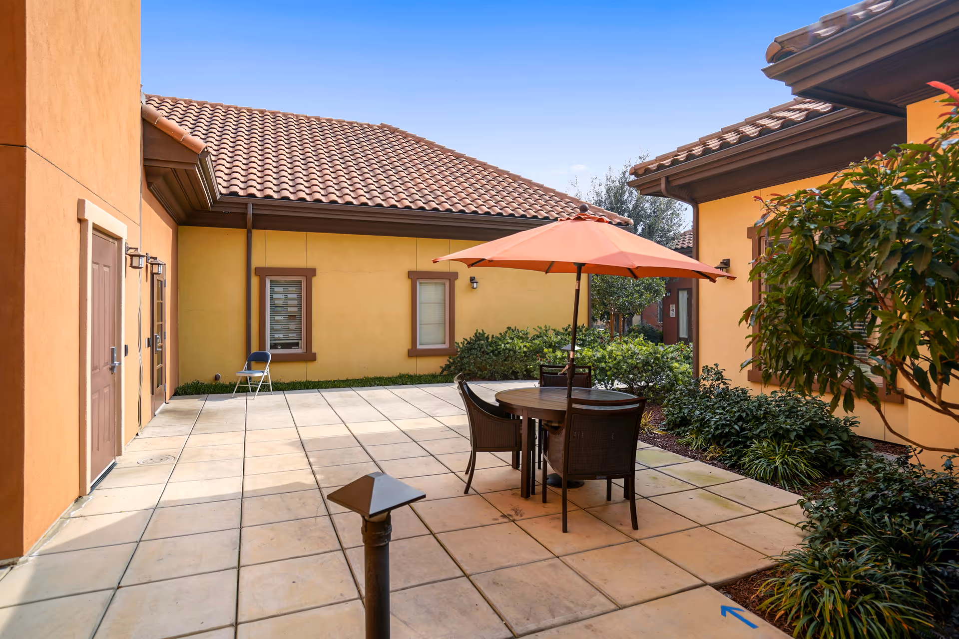 Sunny courtyard patio with a round table and orange umbrella between yellow stucco buildings with tiled roofs.