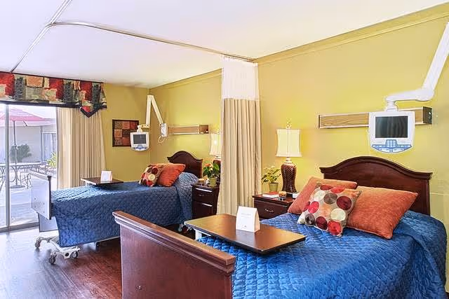A bright and clean healthcare facility room with two hospital-style beds separated by a privacy curtain. Each bed has a blue quilted bedspread and multiple decorative pillows. There are bedside tables with lamps and plants next to each bed. Mounted monitors are positioned above each bed. A sliding glass door leads to an outdoor patio area with tables and chairs.