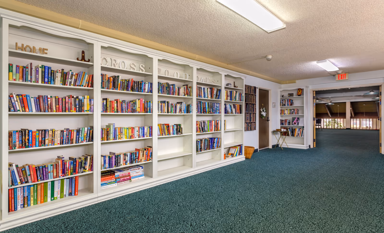 Long carpeted interior hallway with built-in white bookshelves filled with books and an open doorway to a common area.