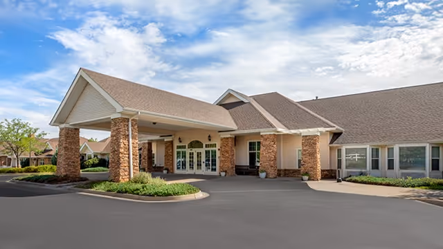 Front entrance of a single-story senior living facility with a covered porte-cochere, stone columns, and a paved driveway under a partly cloudy sky.