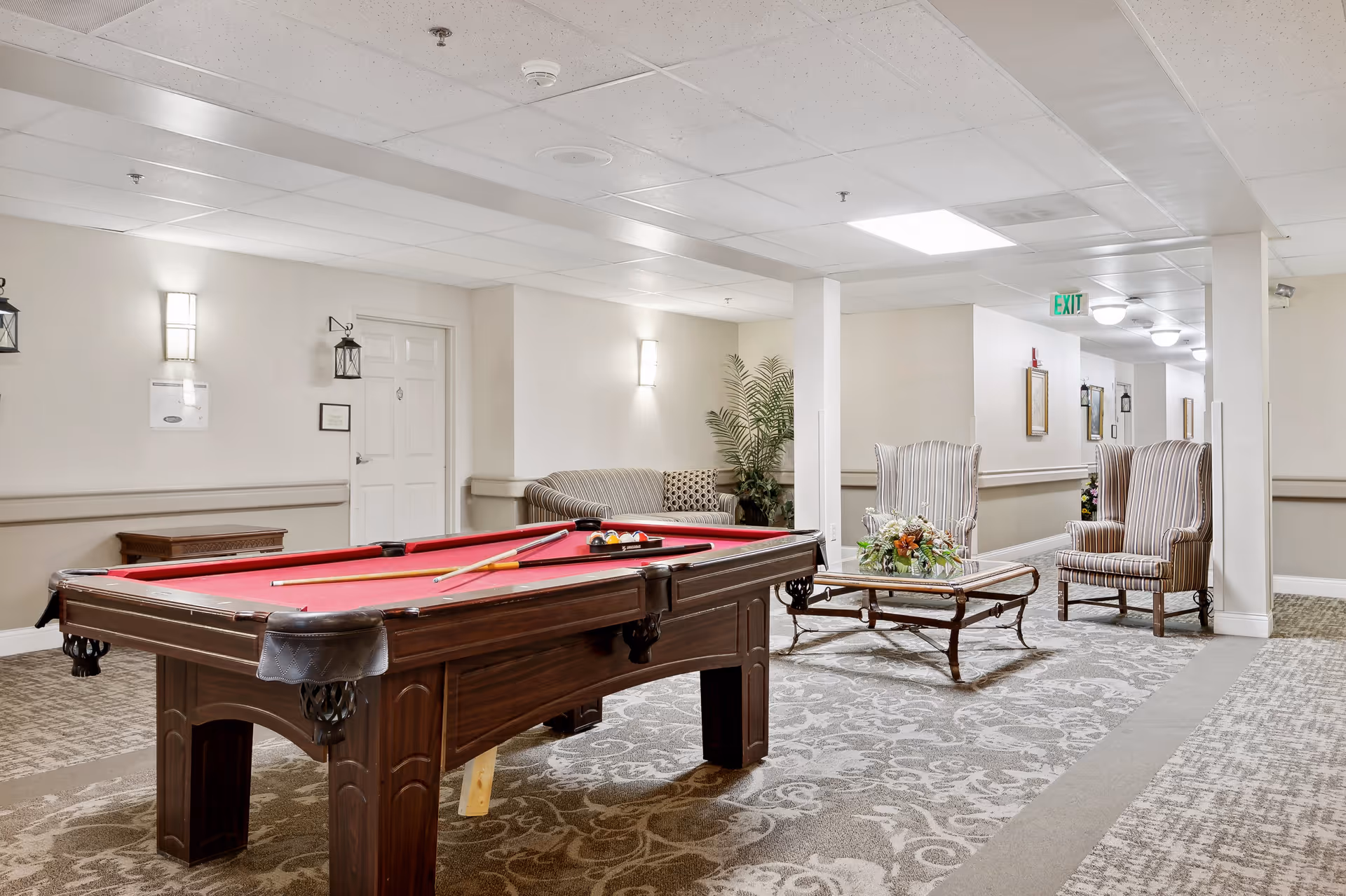 Interior common area of a senior living facility featuring a pool table with red felt, two striped armchairs, a glass coffee table with a floral arrangement, a couch with patterned cushions, and soft lighting on the walls.