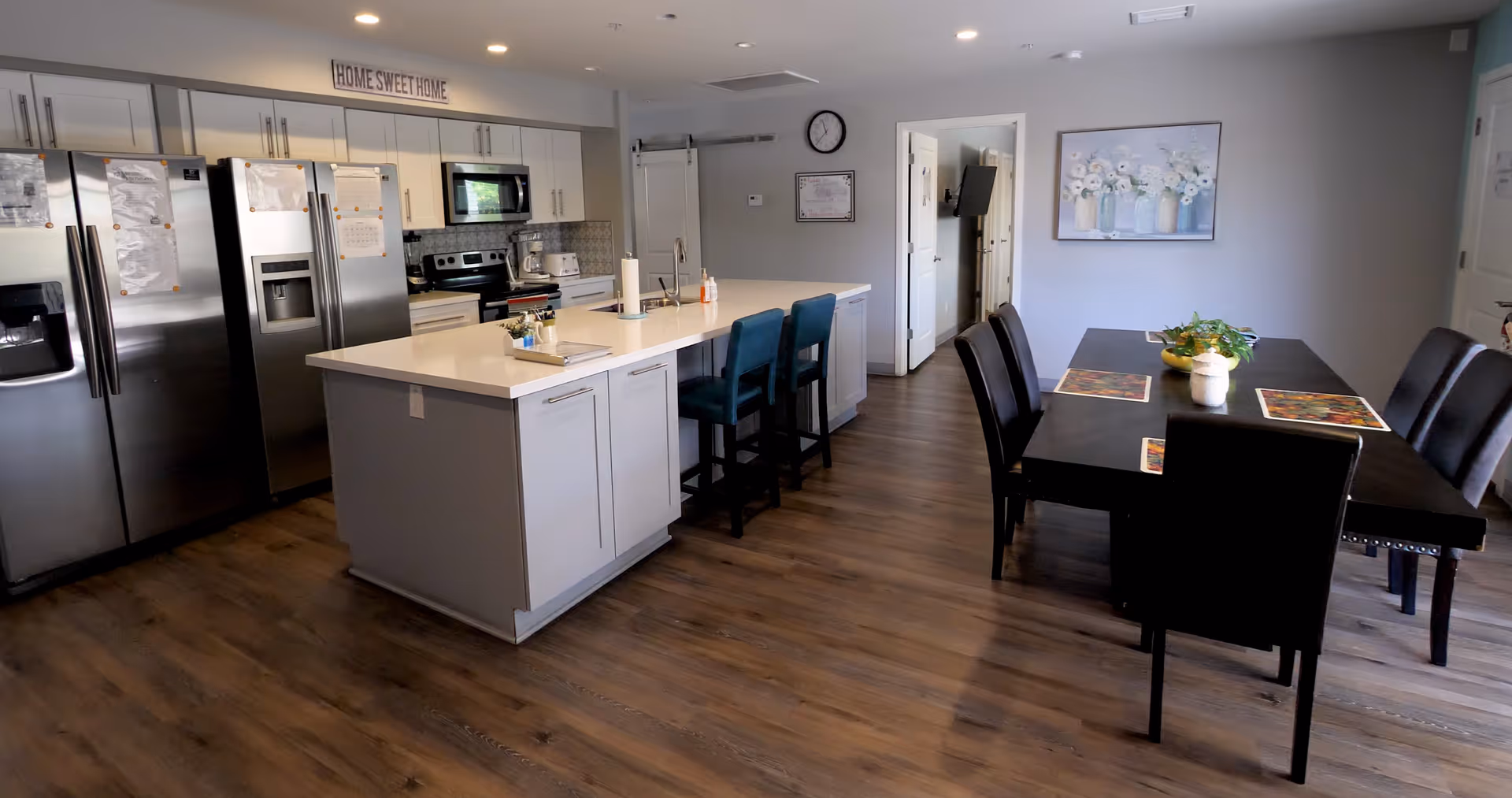 A modern kitchen and dining area with wooden flooring. The kitchen features stainless steel appliances including two refrigerators, a microwave, and an oven. There is a white island with two blue chairs and various kitchen items on the counter. The dining area has a dark wooden table with six black chairs, placemats, and a small plant centerpiece. A painting of white flowers in vases hangs on the wall, and a clock and certificate are visible near an open doorway.