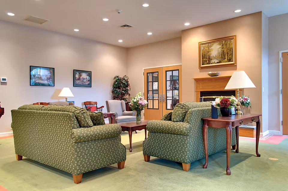 A bright senior living common room with two green patterned sofas facing a coffee table, side tables with lamps and floral arrangements, and framed art on the walls.