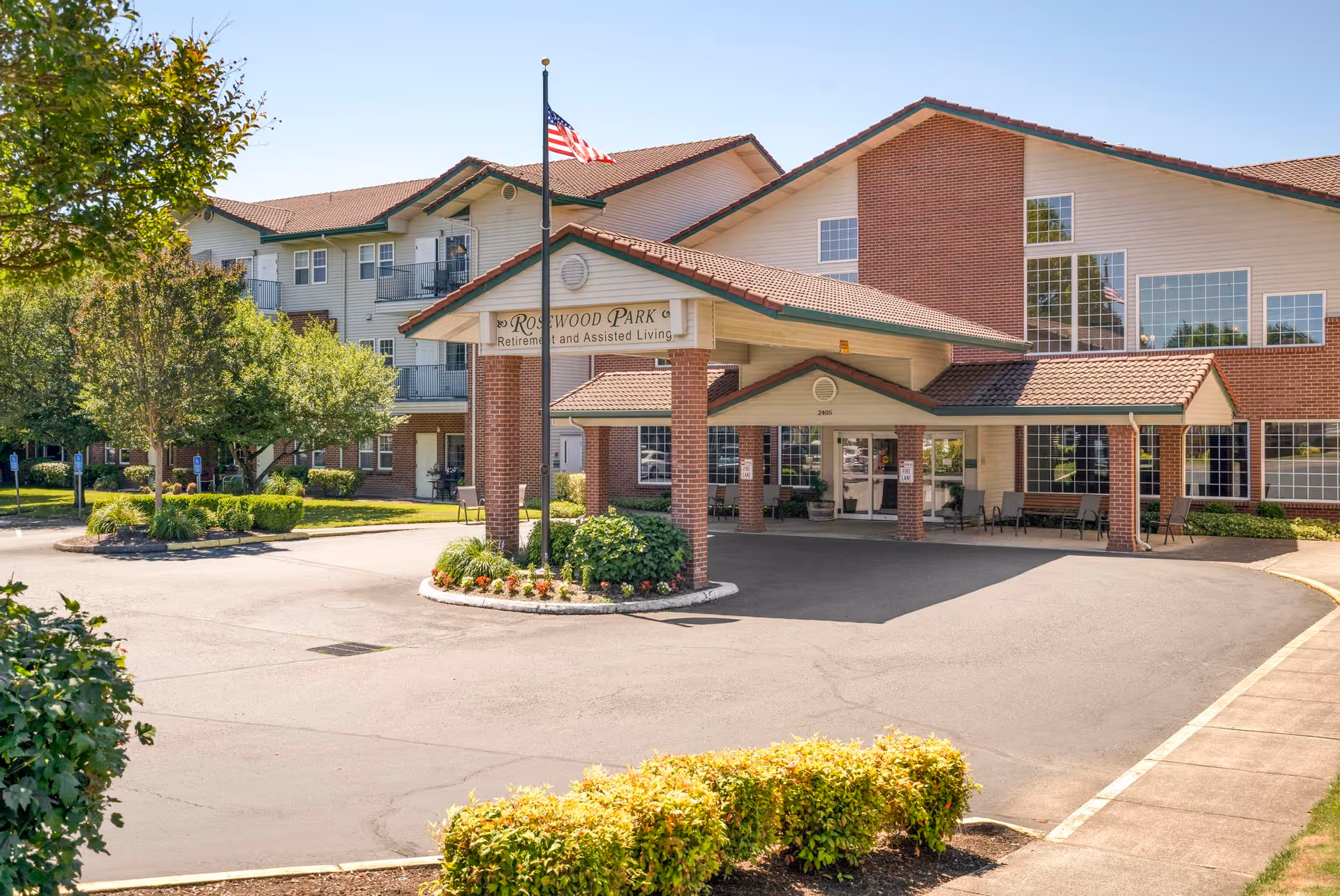 Front entrance of Rosewood Park retirement and assisted living facility with a covered porte-cochere, American flag, and landscaped driveway.