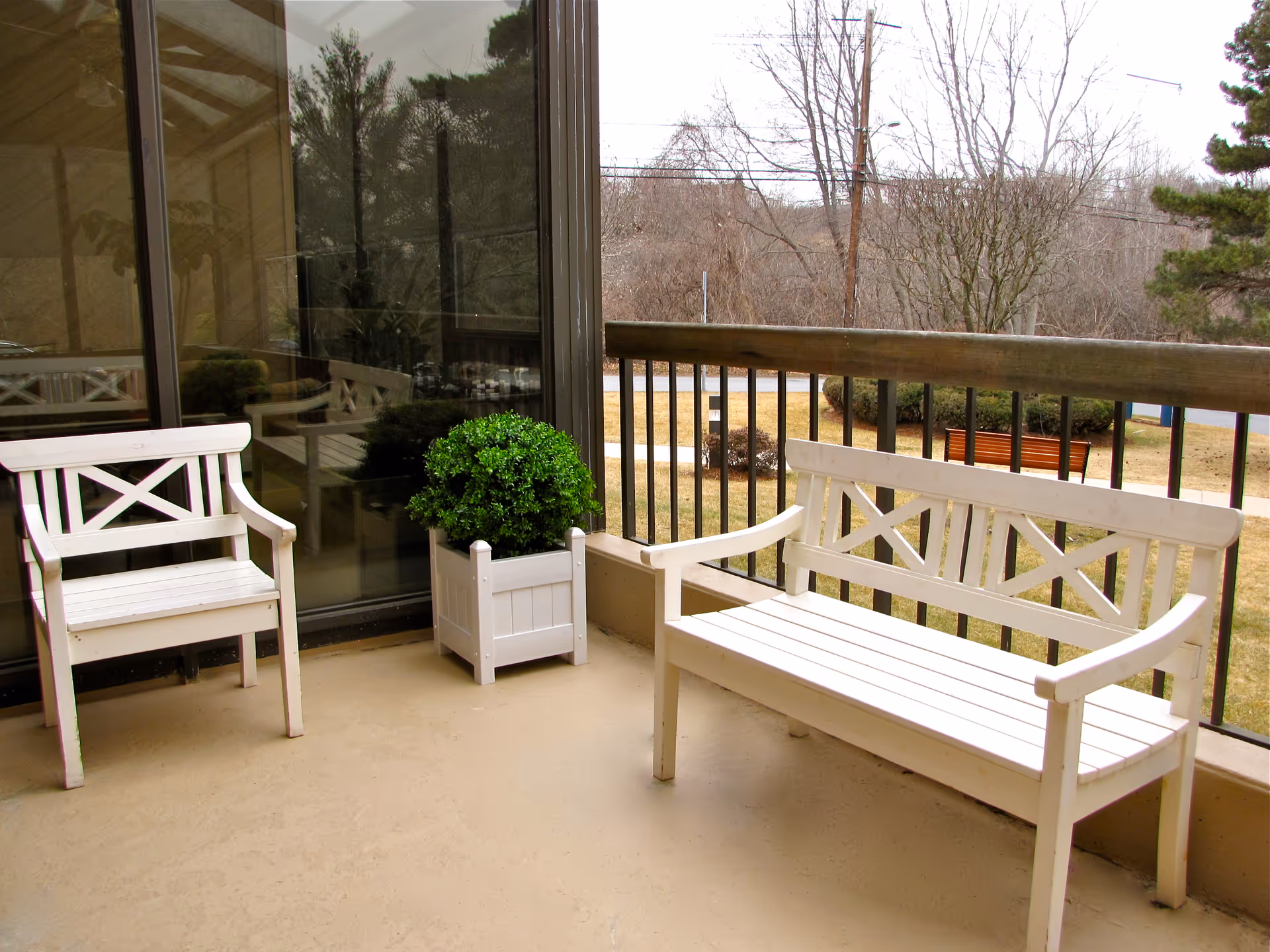 Outdoor balcony area with two white wooden benches and a white planter box containing a green bush. The balcony has a railing and overlooks a grassy area with trees and a bench in the distance.