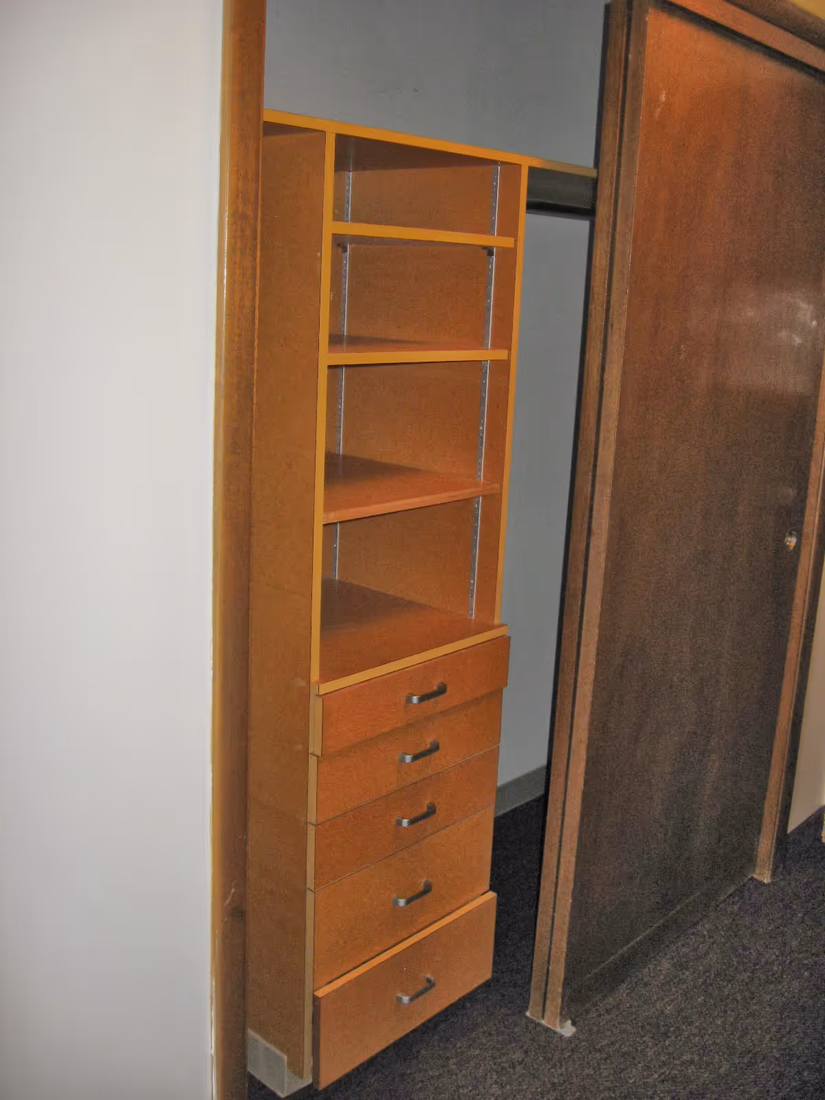 A built-in wooden shelving unit with four shelves and five drawers, located inside a closet with a sliding wooden door partially open, showing a carpeted floor and plain walls.