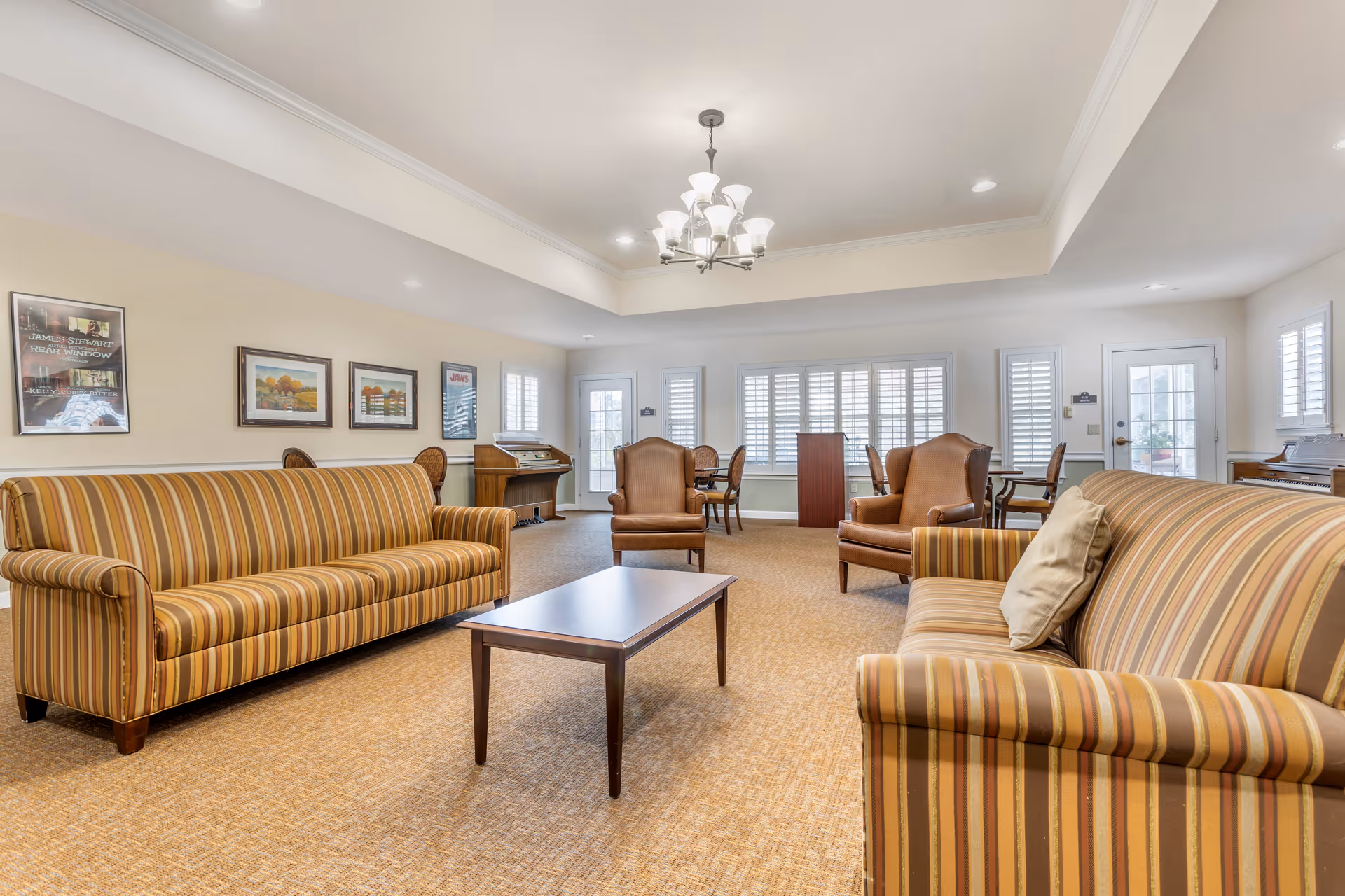 A spacious living room with two striped sofas facing each other and a wooden coffee table in between. Two brown armchairs are positioned near the back wall with large windows covered by white shutters. The room has beige walls adorned with framed pictures and a chandelier hanging from the ceiling. There is a piano and a jukebox near the back corners of the room.