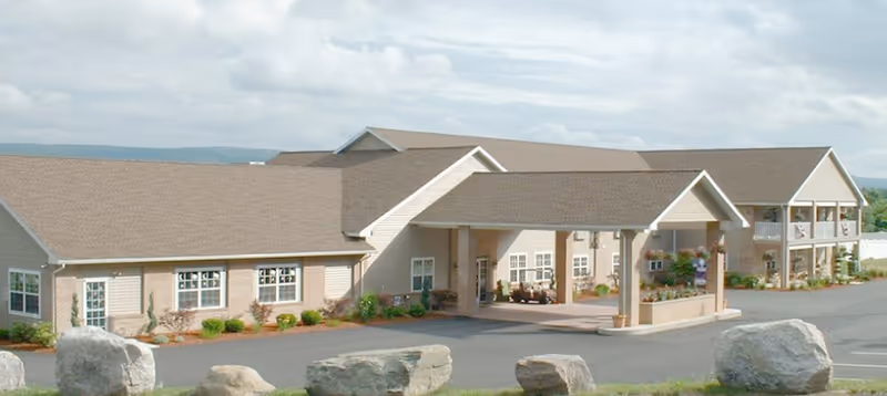 Exterior view of a single-story senior living facility building with beige siding and a covered entrance. The building is surrounded by a paved driveway and landscaped with shrubs and large rocks in the foreground. The sky is partly cloudy.