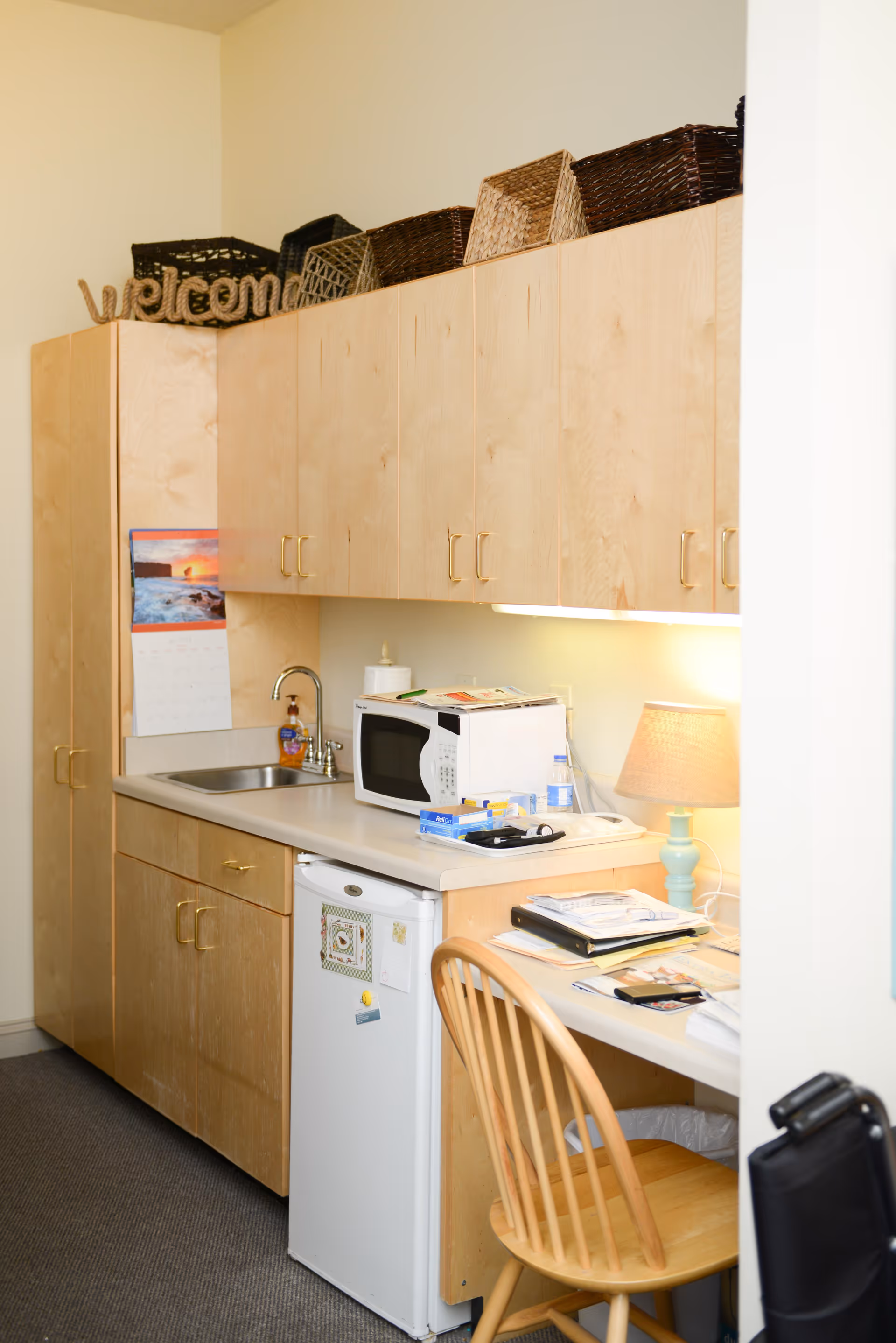 A small kitchenette area with light wood cabinets, a countertop with a microwave, a small sink, and a mini refrigerator. There are several woven baskets on top of the cabinets and a decorative 'welcome' sign. A wooden chair is placed at the counter, which has various papers and a lamp with a light blue base. A calendar with a sunset image is hanging on the side of the cabinet.