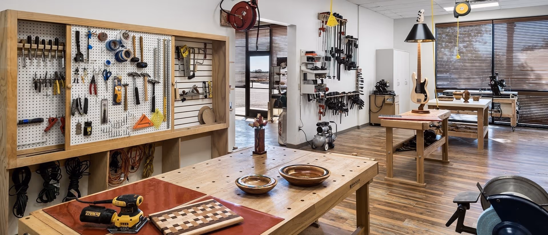 Well-lit woodworking and craft workshop with wooden workbenches, pegboards of tools, power tools and large windows.