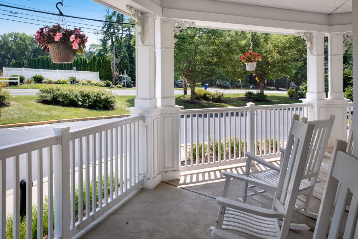 A covered porch area with white wooden railings and several white rocking chairs. Two hanging flower pots with pink and red flowers are suspended from the porch ceiling. The porch overlooks a landscaped area with green bushes, trees, and a road in the background.